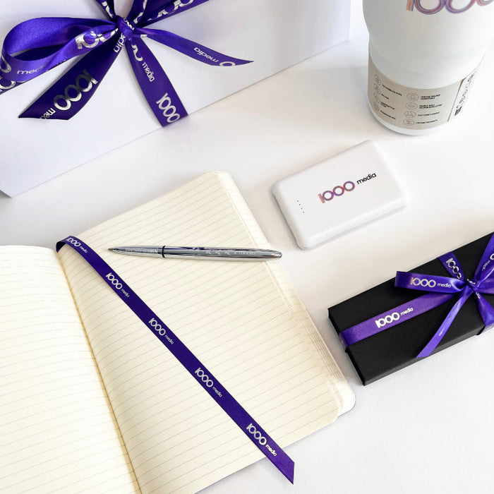 Notebook, pen, and gift box with a purple ribbon on a white surface, featuring the 1000 Media logo.