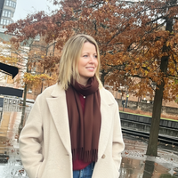 Women wearing brown scarf & beige coat with trees with autumn leaves on.