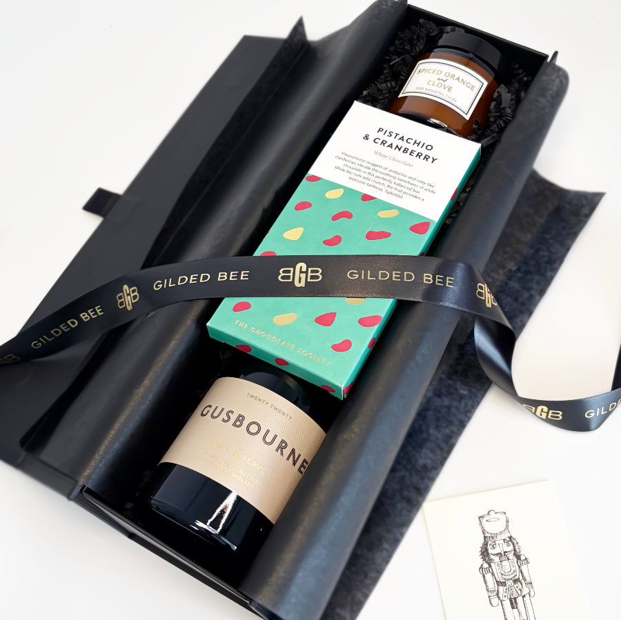 A curated gift box set from Gilded Bee, featuring a black candle, a boxed pistachio & cranberry spread, and a bottle of Gusbourne cheese, wrapped in a branded black ribbon.