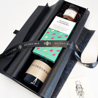 A curated gift box set from Gilded Bee, featuring a black candle, a boxed pistachio & cranberry spread, and a bottle of Gusbourne cheese, wrapped in a branded black ribbon.