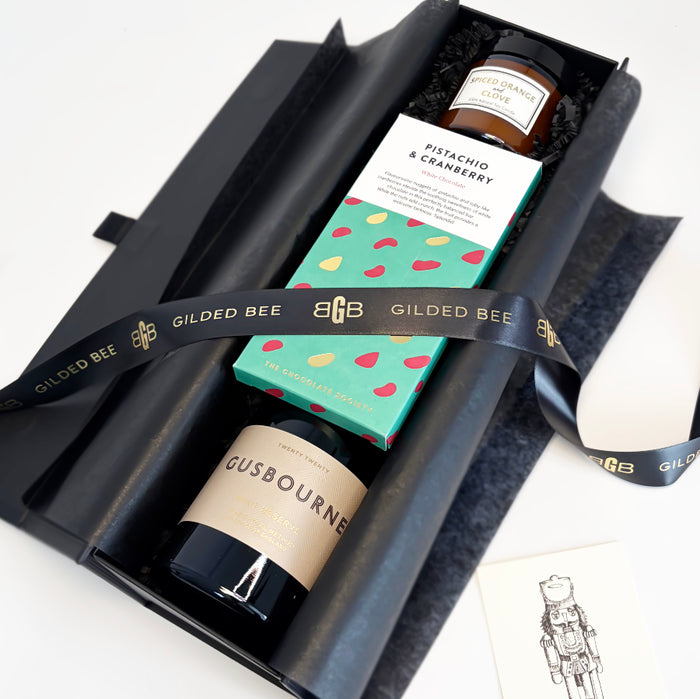 A curated gift box set from Gilded Bee, featuring a black candle, a boxed pistachio & cranberry spread, and a bottle of Gusbourne cheese, wrapped in a branded black ribbon.