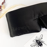 Black sleep mask with 'Drowsy' branding on a white surface with a bee illustration.