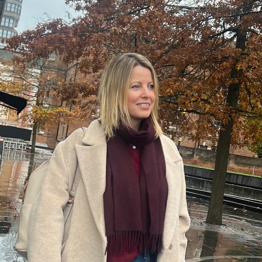 Woman wearing a beige coat and burgundy scarf standing outdoors with autumn trees in the background