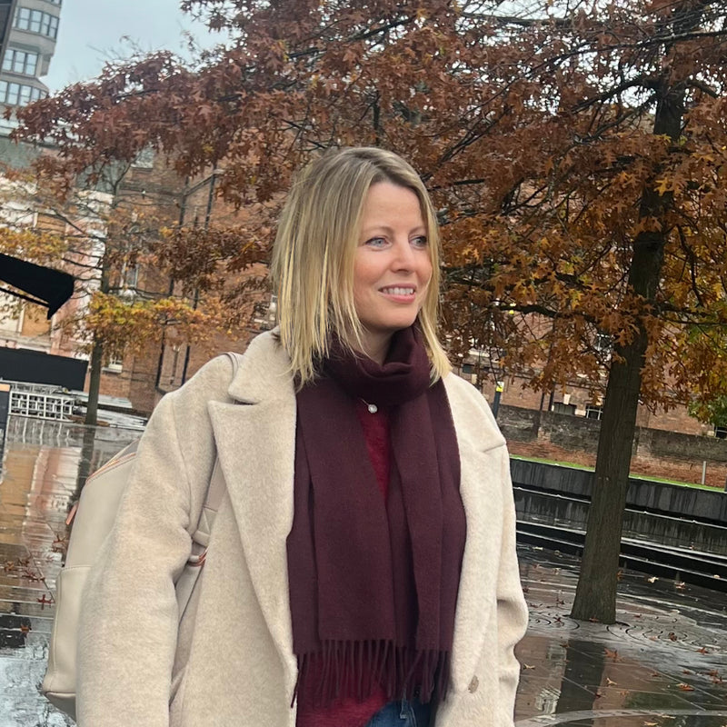 Woman wearing a beige coat and burgundy scarf standing outdoors with autumn trees in the background
