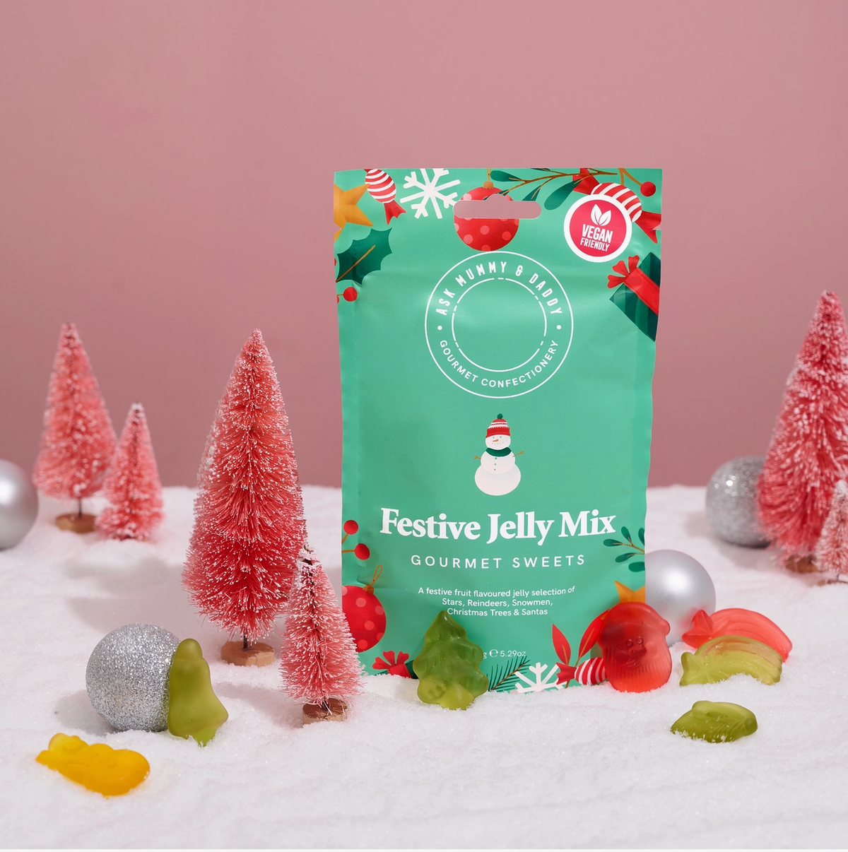 A green bag of 'Festive Jelly Mix' gourmet sweets by The Flower & Tree stands upright on a white snowy surface decorated with miniature pink Christmas trees and candy canes, against a solid pink background.
