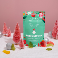 A green bag of 'Festive Jelly Mix' gourmet sweets by The Flower & Tree stands upright on a white snowy surface decorated with miniature pink Christmas trees and candy canes, against a solid pink background.