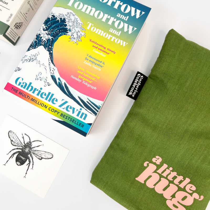Book titled 'Tomorrow and Tomorrow and Tomorrow' by Gabrielle Zevin with a green hot water bottle with the words a little hug on a white surface.