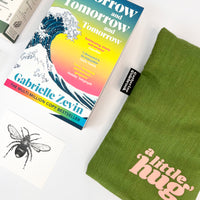 Book titled 'Tomorrow and Tomorrow and Tomorrow' by Gabrielle Zevin with a green hot water bottle with the words a little hug on a white surface.
