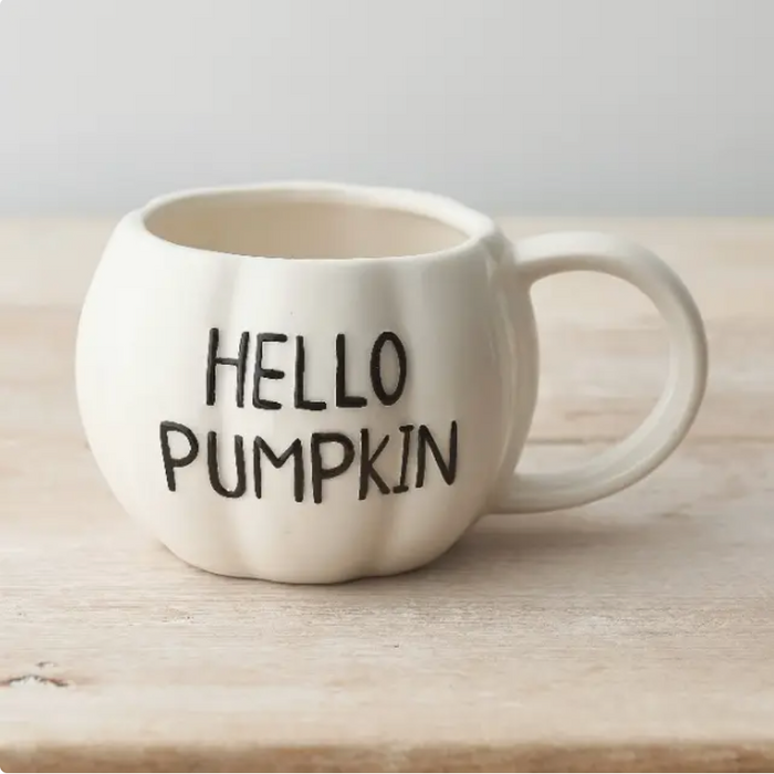 An off-white ceramic mug shaped like a pumpkin with the words 'HELLO PUMPKIN' printed in black on the front, resting on a light wooden surface.
