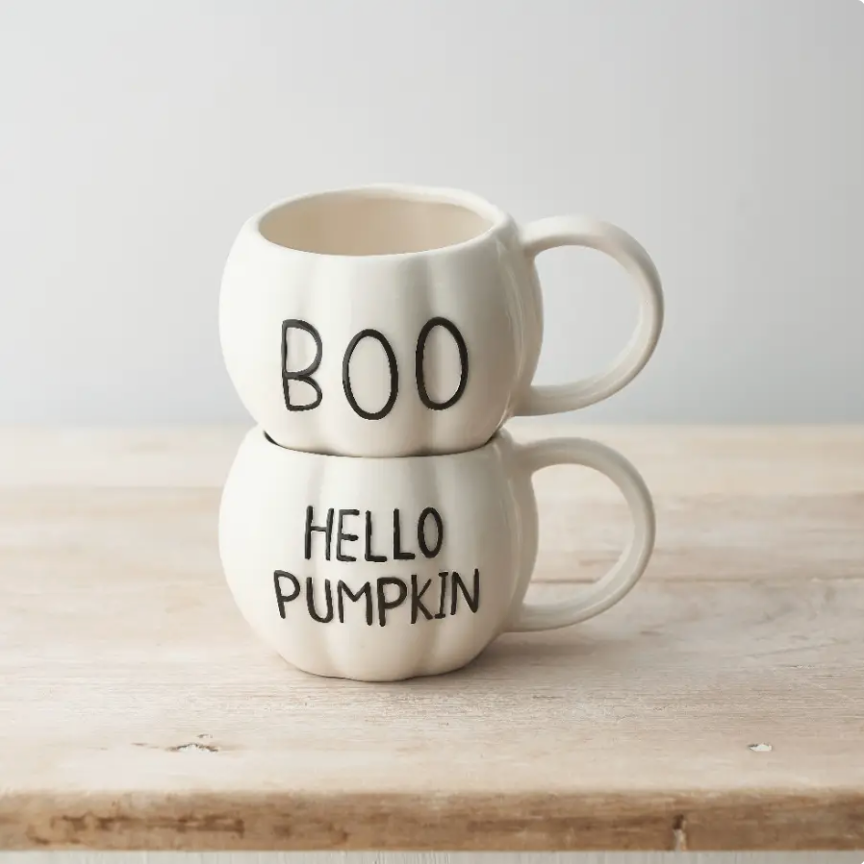 Two stacked ceramic mugs shaped like pumpkins on a light wood surface against a soft light gray background. The top mug says "BOO" in black lettering, and the bottom mug says "HELLO PUMPKIN" in black lettering.