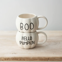 Two stacked ceramic mugs shaped like pumpkins on a light wood surface against a soft light gray background. The top mug says "BOO" in black lettering, and the bottom mug says "HELLO PUMPKIN" in black lettering.