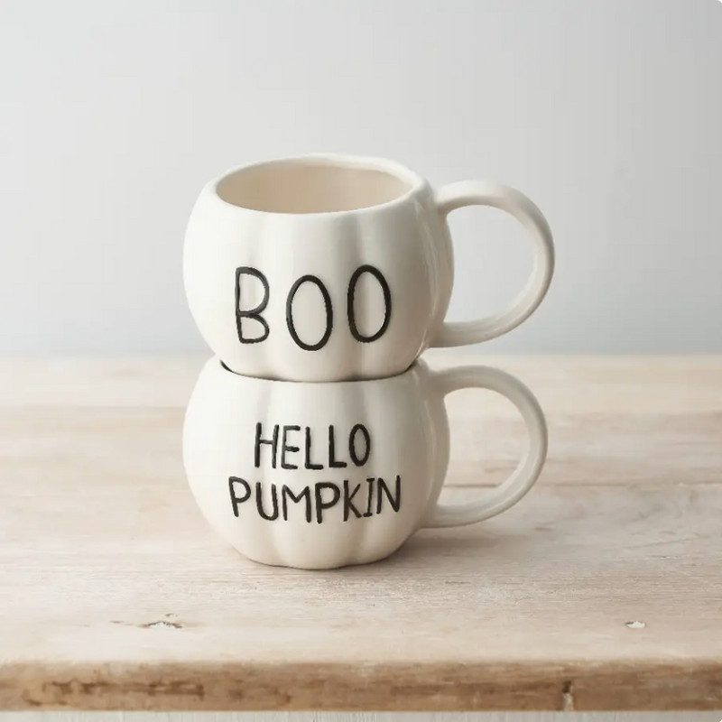 Two stacked ceramic mugs shaped like pumpkins on a light wood surface against a soft light gray background. The top mug says "BOO" in black lettering, and the bottom mug says "HELLO PUMPKIN" in black lettering.