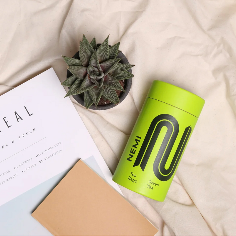 Green NEMI tea bag container with a plant and notebook on a light surface