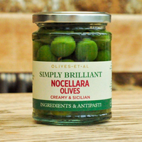 A glass jar of Simply Brilliant Nocellara Olives on a wooden surface with a blurred rustic background. The jar contains green olives in a brine with a white, green, and red label.