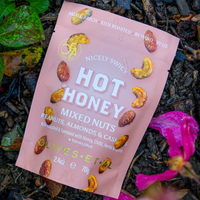 A pink bag of Nicely Swicy Hot Honey Mixed Nuts lies on dark mulch with green leaves and a bright pink flower nearby. The bag features white text and illustrations of roasted nuts.
