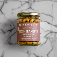 A glass jar of Olives Et Al Pigs in Blankets mixed nuts, featuring a dusty rose label and gold lid, set against a white and gray marble background.