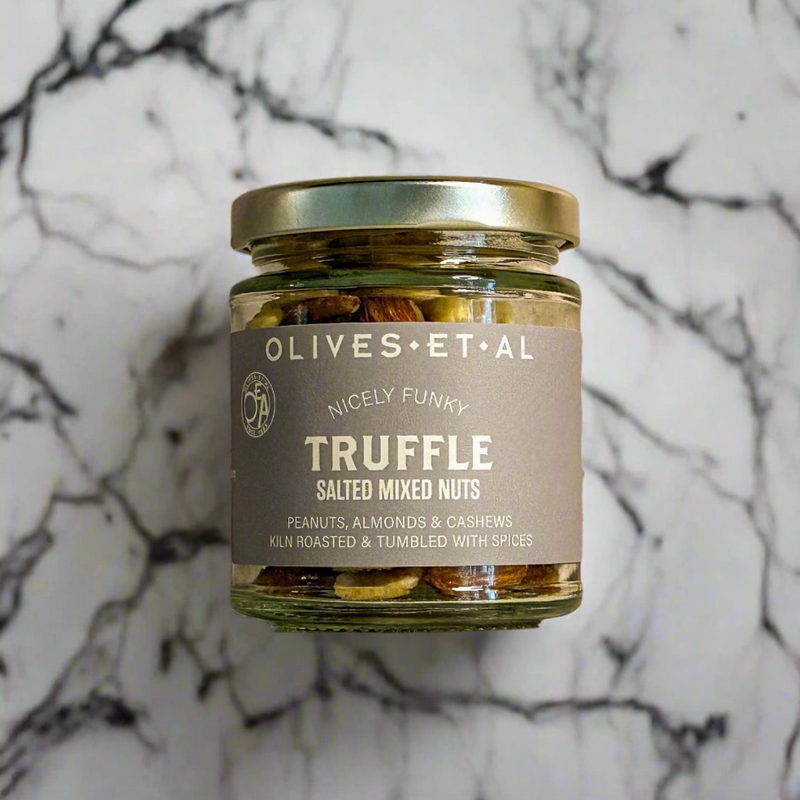 A glass jar of Olives Et Al Truffle Salted Mixed Nuts with a gold lid and a brownish-gray label, set against a white marble background with black veining.