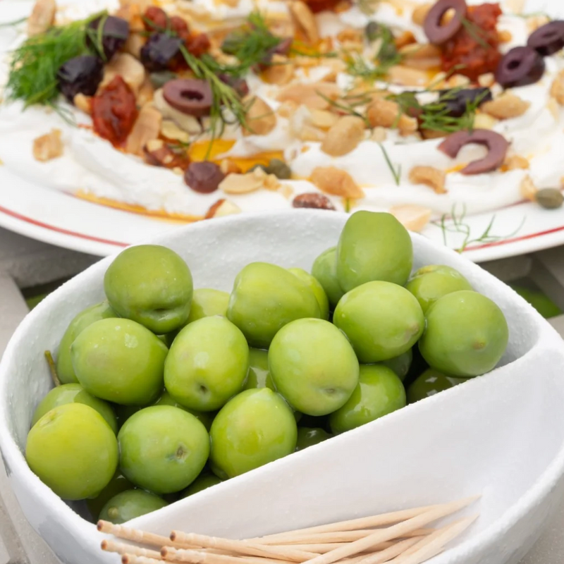 A white divided bowl filled with green plums in the foreground, with wooden toothpicks in the lower section and a plated appetizer dish blurred in the background.