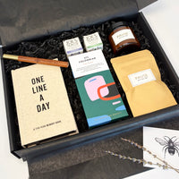 A curated gift box set displayed inside an open black box with black shredded filler. Visible items include a beige hardcover book titled 'ONE LINE A DAY', a gold pen, a box of Joe's Tea 'Colombian', a candle jar labeled 'PAUSE', a beige pouch labeled 'PAUSE', and a small card with a bee illustration.