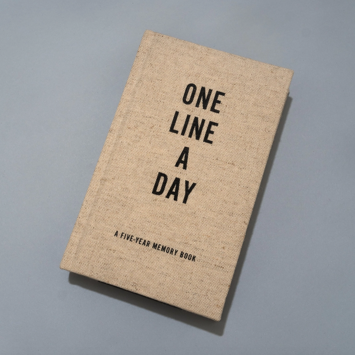 A square-shaped beige linen-bound memory book titled 'ONE LINE A DAY' with black text, photographed on a neutral gray background.