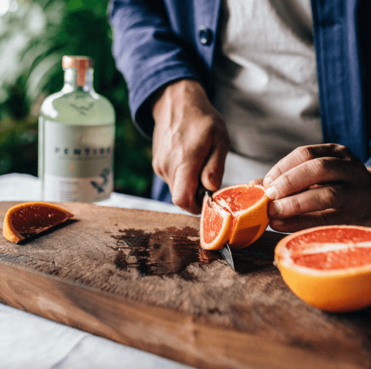 Person cutting a grapefruit on a wooden cutting board with a bottle of gin in the background.