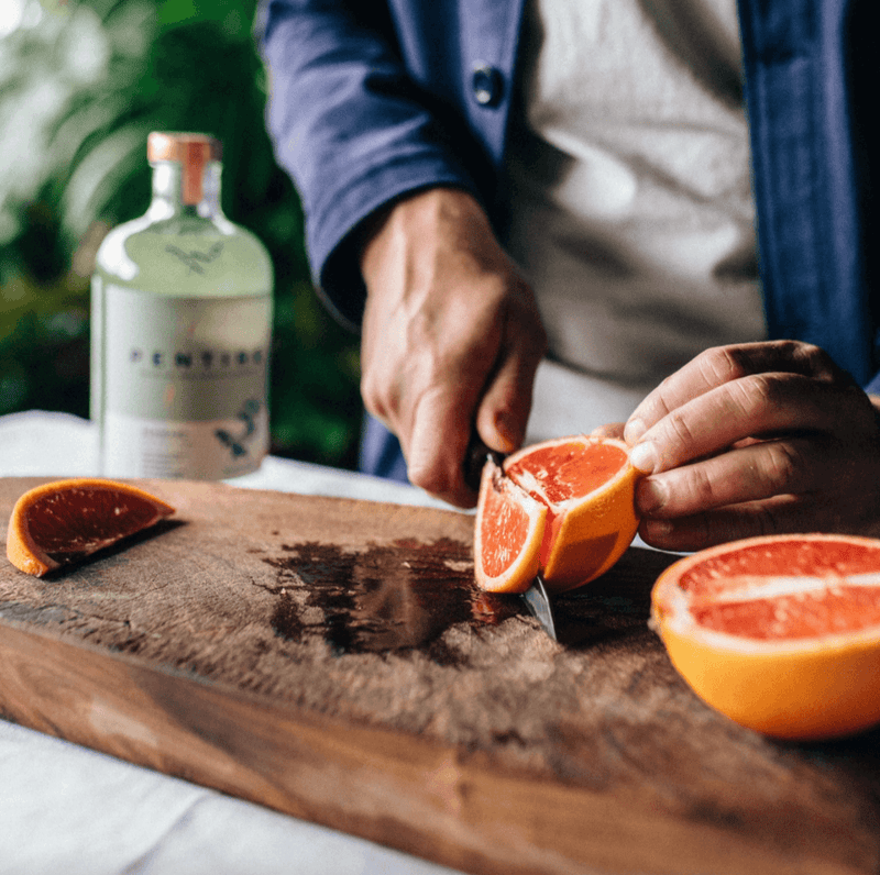 Person cutting a grapefruit on a wooden cutting board with a bottle of gin in the background.