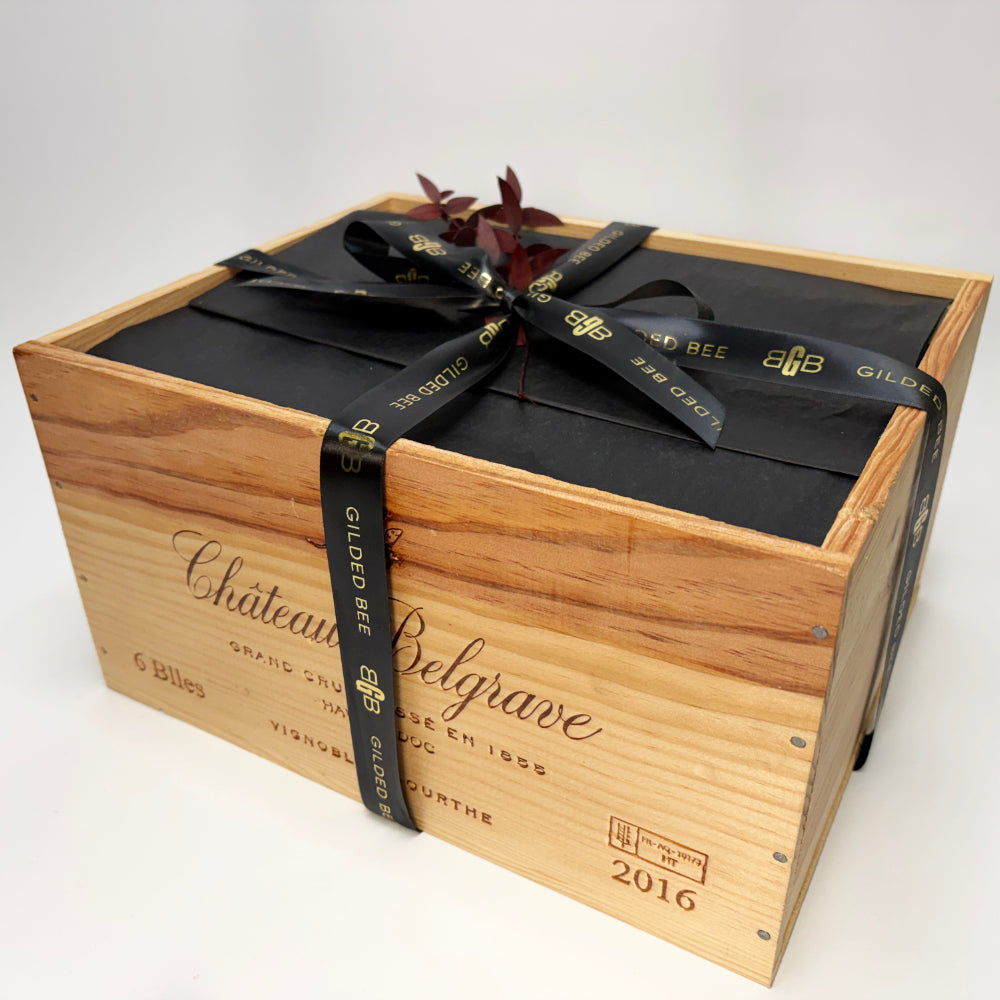 A wooden wine crate with Château Belgrave branding, wrapped in a black gift bag tied with a black ribbon printed with Gildan Bee logos, topped with a small dark leafy sprig, on a white studio background.