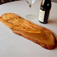 An irregularly shaped olive wood serving board with a natural live edge, displayed on a white table next to a wine bottle and a wine glass.