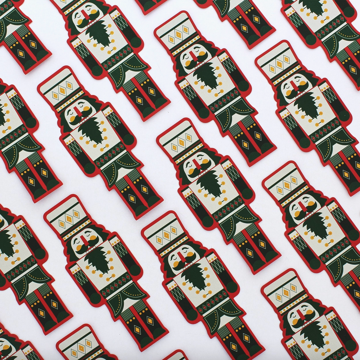 A repeating pattern of nutcracker-shaped items on a white background, each featuring a cream face and torso with dark green and red clothing details, yellow geometric accents, and a black beard.