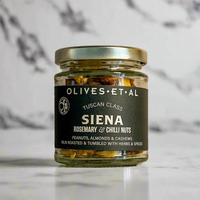 A glass jar of Olives Et Al Siena Tuscan Class roasted nuts with a black label and gold lid, set on a white marble surface against a marble background.