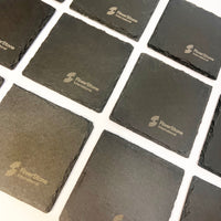 A set of square dark gray RiverStone International slate coasters arranged on a white background.
