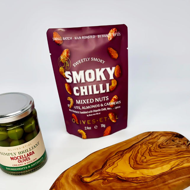 A curated gift set on a white background featuring a jar of Smokey Smoked Nocky Pickles, a pouch of Smokey Smoked Smokey Chilli mixed nuts, a natural olive wood serving board, a green leafy sprig, and a branded card with a bee illustration.