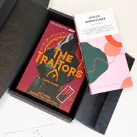 A boxed card game called The Traitors displayed next to a chocolate bar labeled Bitter Marmalade, inside an open black presentation box on a white surface.