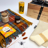 A curated gift set arranged on a white surface, featuring a multicolor plaid fringed scarf, a cream knit sweater with a 'Happy Knitting' band, a wooden wine box, a yellow cylindrical tin, a jar of bath salts, a candle, and a Love Cocoa box.