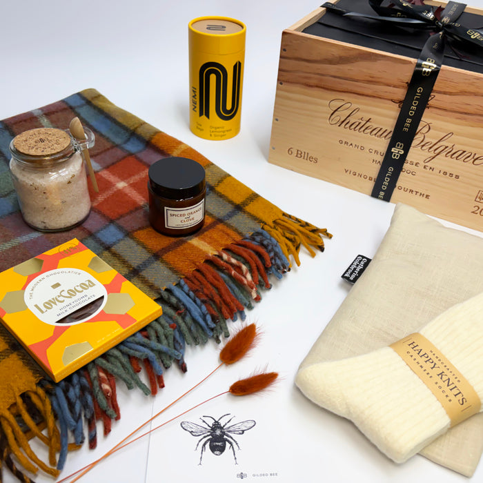 A curated gift set arranged on a white surface, featuring a multicolor plaid fringed scarf, a cream knit sweater with a 'Happy Knitting' band, a wooden wine box, a yellow cylindrical tin, a jar of bath salts, a candle, and a Love Cocoa box.
