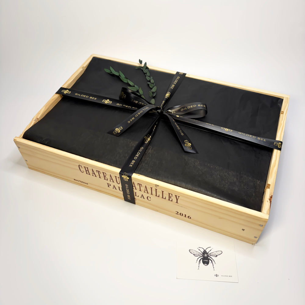 A black-wrapped gift box presented in a light natural wood wine crate, tied with a black ribbon printed with gold lettering, on a clean white background. A small bee card is placed in front.