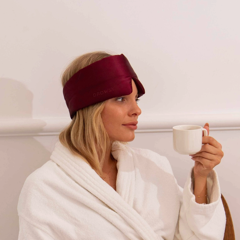 Woman wearing a burgundy satin eye mask and holding a white mug, wrapped in a white robe.