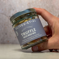 A hand holding a glass jar of Olives Et Al Truffle salted mixed nuts against a neutral, softly blurred background.