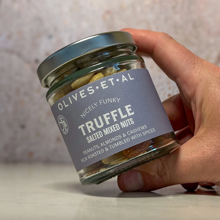 A hand holding a glass jar of Olives Et Al Truffle salted mixed nuts against a neutral, softly blurred background.
